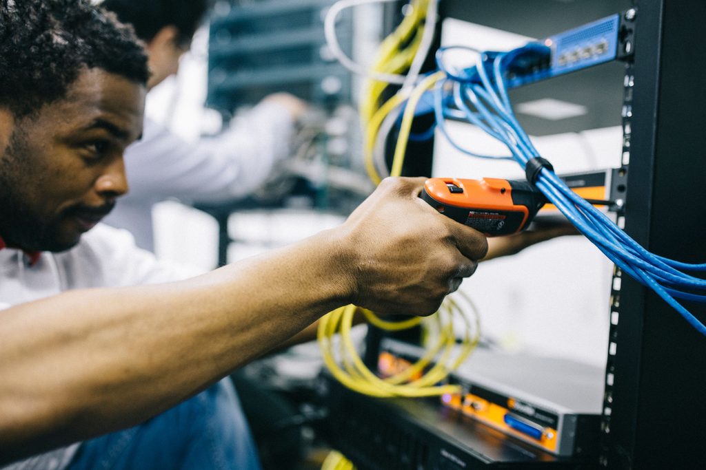 An engineer plugging a network cable into a patch panel, a colleague working behind
