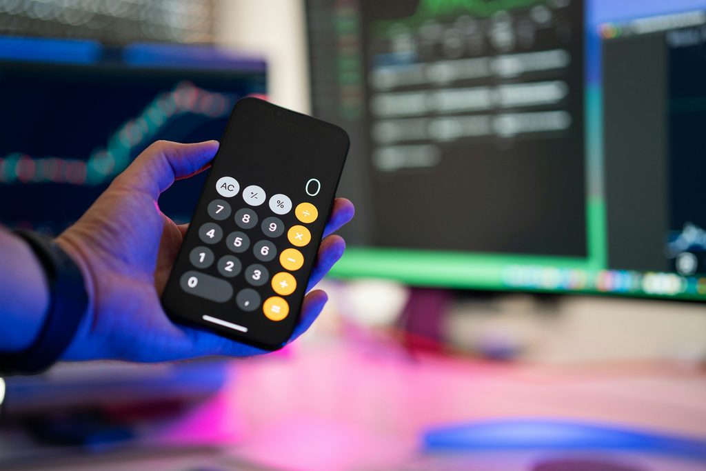 A hand holding a calculator in front of brightly lit computer screens