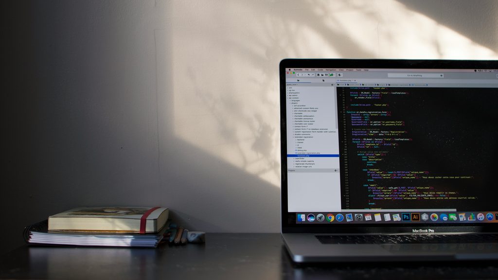 A laptop displaying code on a dark desk, next to a closed notebook and pencils