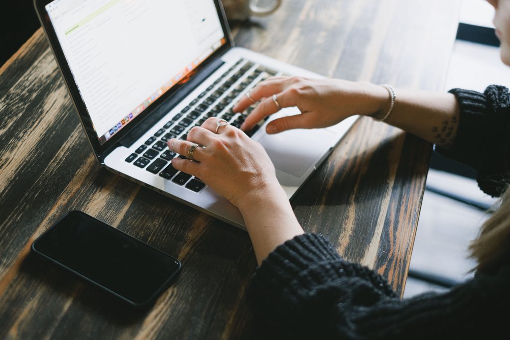 Les mains d’une femme sur le clavier d’un ordinateur portable posé sur une table en bois, un téléphone à côté
