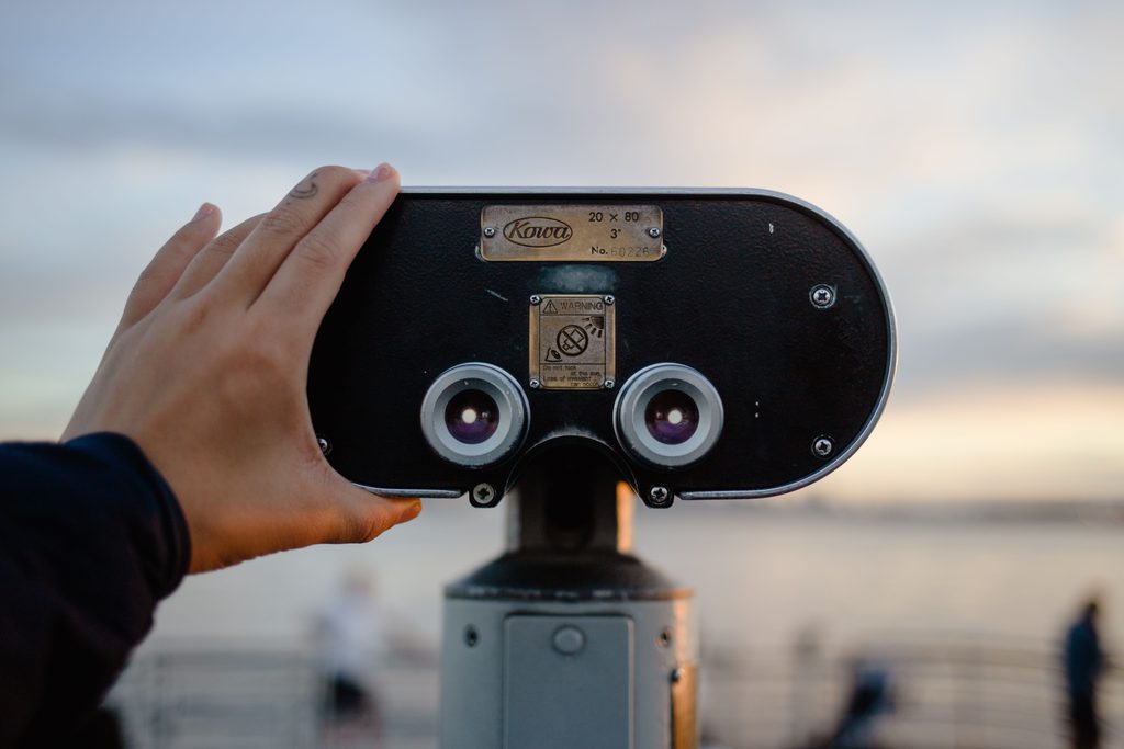 Deux mains tiennent une longue-vue panoramique orientée vers la mer, au crépuscule