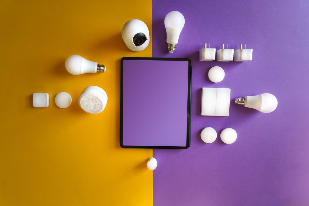Vue de dessus d’objets connectés blancs — ampoules, prises, caméra — disposés autour d’une tablette, sur fond jaune et violet