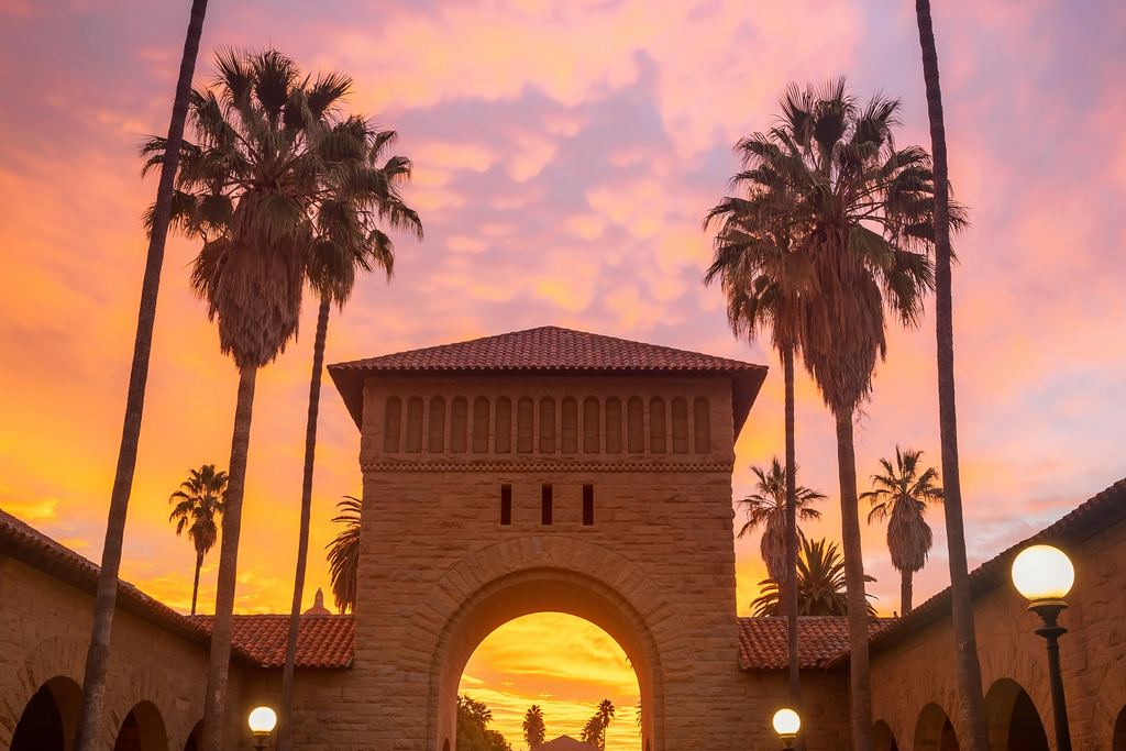 Le portail d’entrée du campus de Stanford, encadré de palmiers, sous un ciel de crépuscule orangé