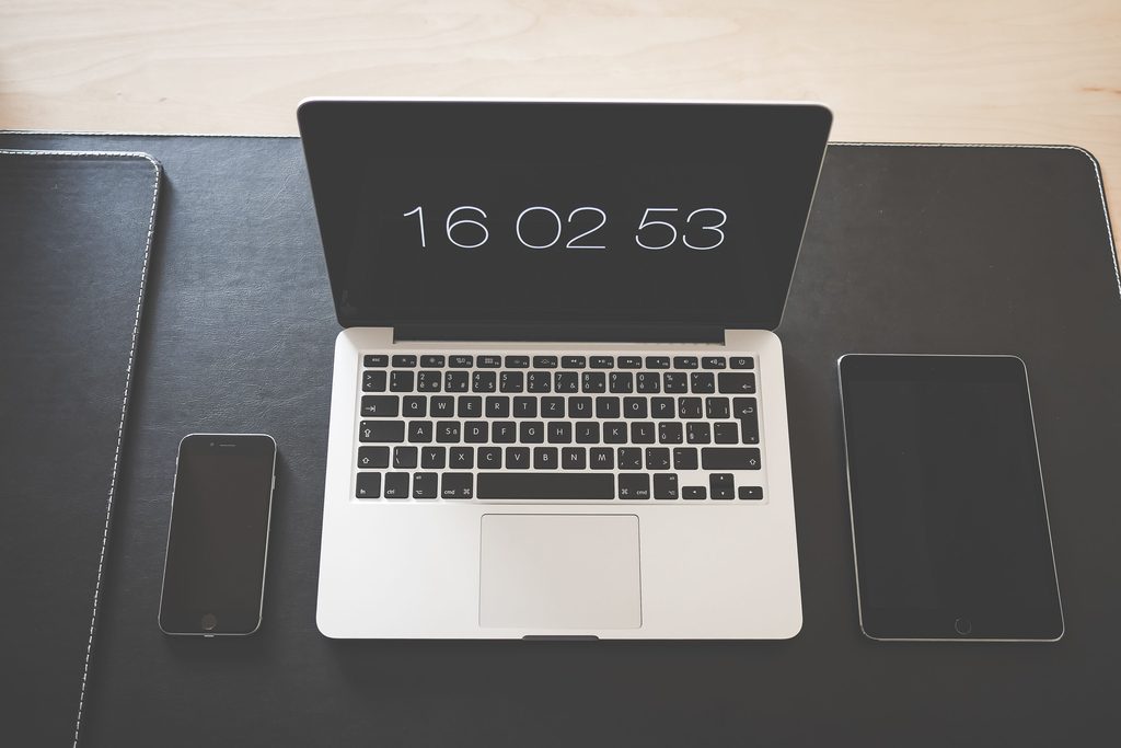 A desk seen from above: an open laptop, a phone and a tablet on a desk mat