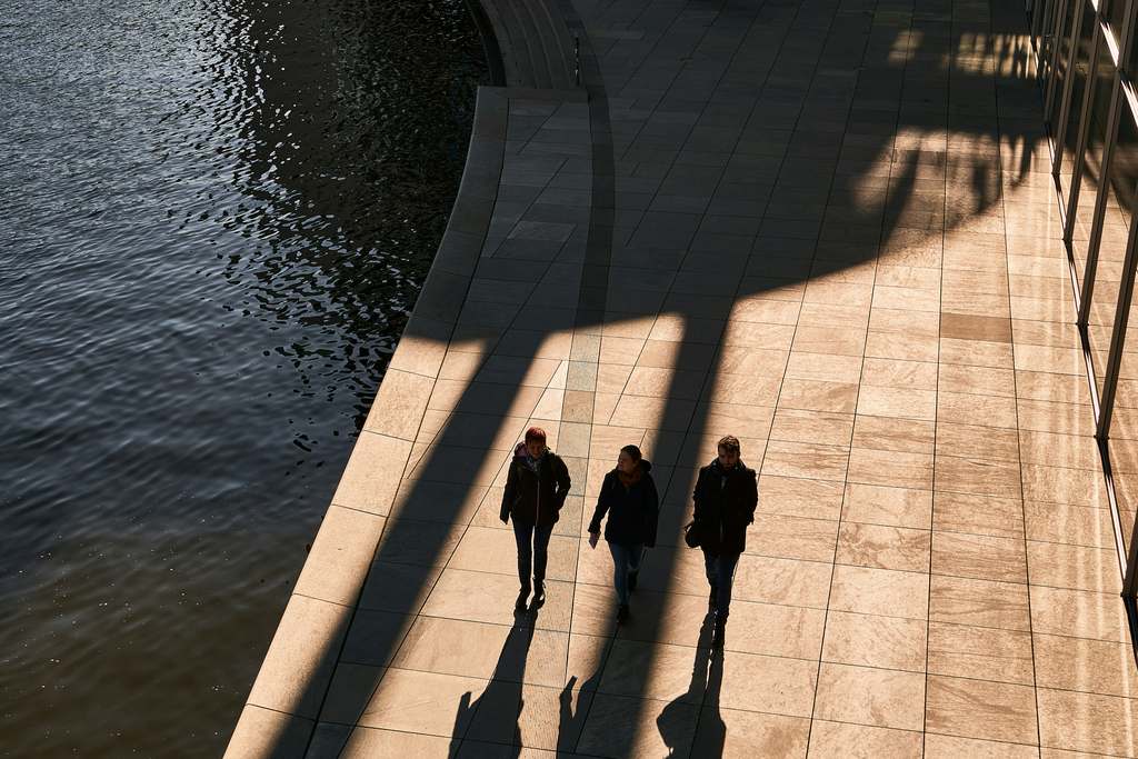 Trois silhouettes marchent sur un quai au bord de l’eau, vues de haut, ombres allongées
