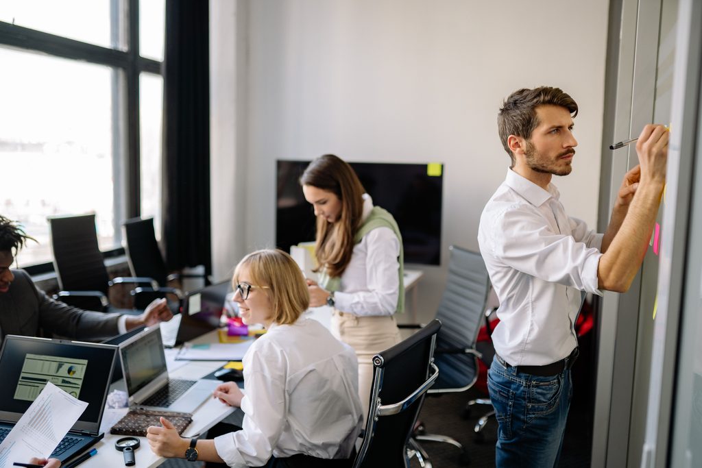 Dans un bureau ouvert, un homme écrit sur un tableau blanc pendant que deux collègues travaillent
